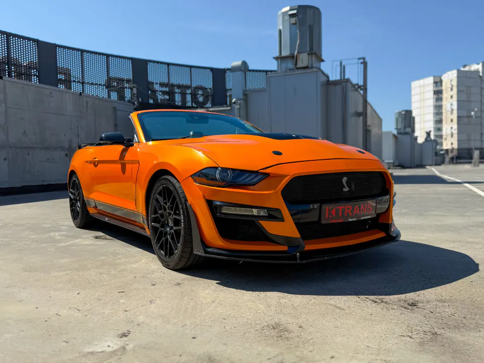 Orange 2020 Ford Mustang convertible with a KTRANS plate on a parking garage roof, city buildings behind