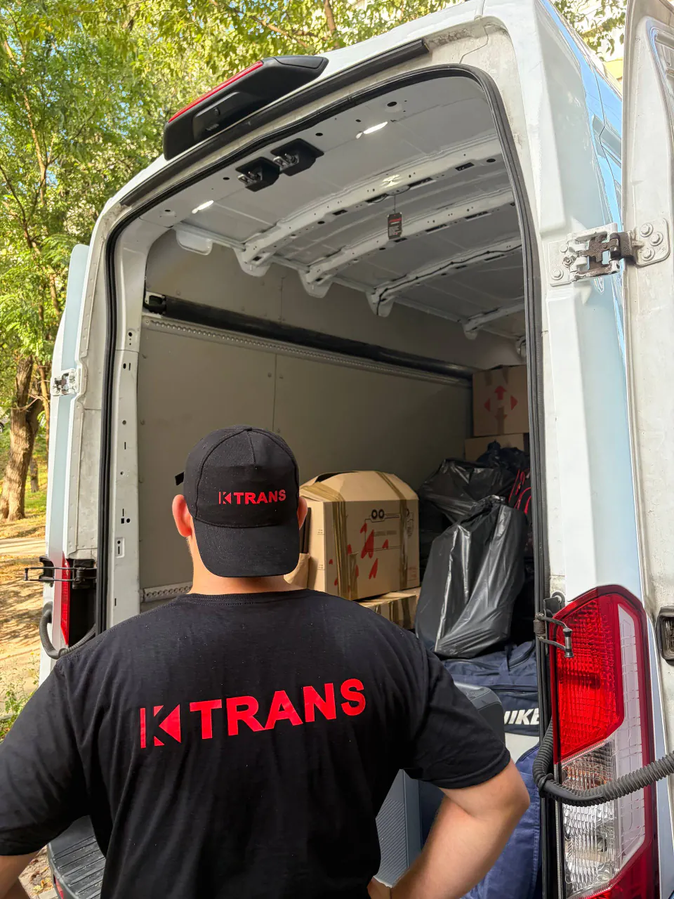 Employee in a KTRANS cap and T-shirt loading boxes into a van, seen from behind